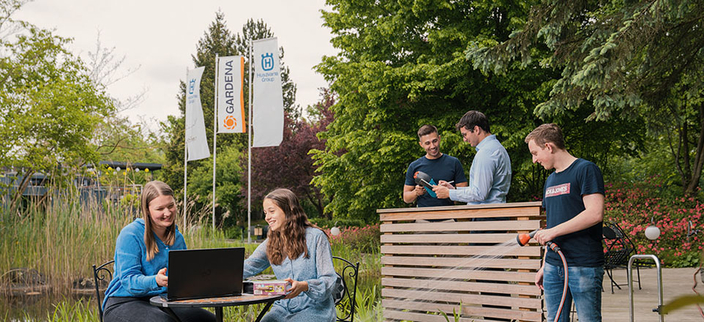 Fünf junge Personen auf einem Gartengelände mit kleinem Teich, zwei sitzen am Laptop, einer bewässert den Garten, zwei sind im Gespräch Fünf junge Personen auf einem Gartengelände mit kleinem Teich, zwei sitzen am Laptop, einer bewässert den Garten, zwei sind im Gespräch