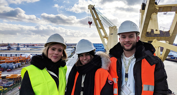 Drei junge lächelnde Personen mit Warnweste und Helm am Hafen Drei junge lächelnde Personen mit Warnweste und Helm am Hafen