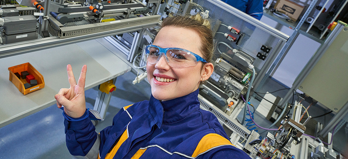 Auszubildende mit Schutzbrille zeigt mit den Fingern ein V 