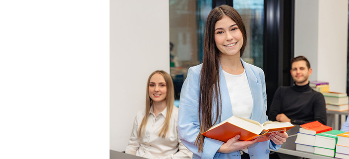 Eine Person hält ein Buch in der Hand und lächelt in die Kamera. Auf dem Tisch hinter ihr liegen weitere Bücher. Zwei Personen sitzen im Hintergrund mit Blick zur Kamera. Eine Person hält ein Buch in der Hand und lächelt in die Kamera. Auf dem Tisch hinter ihr liegen weitere Bücher. Zwei Personen sitzen im Hintergrund mit Blick zur Kamera.