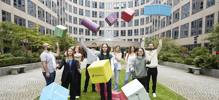Gruppe von neun jungen Personen wirft bunte Sitzwürfel in die Luft vor dem Gebäude der GEWOBAG