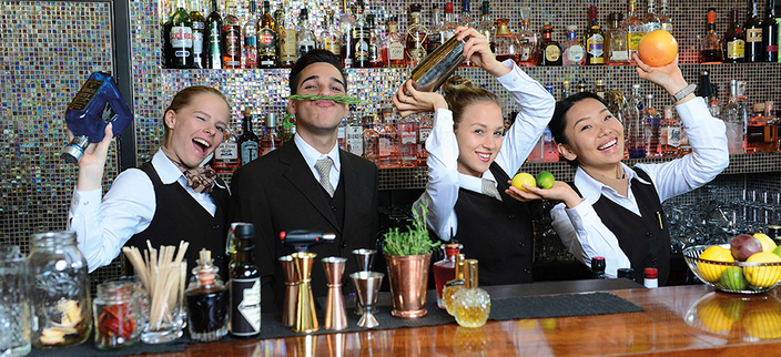 Vier Personen stehen in eleganter Kleidung hinter einer Bar, machen Spaß und halten verschiedene Barutensilien und Früchte in den Händen.