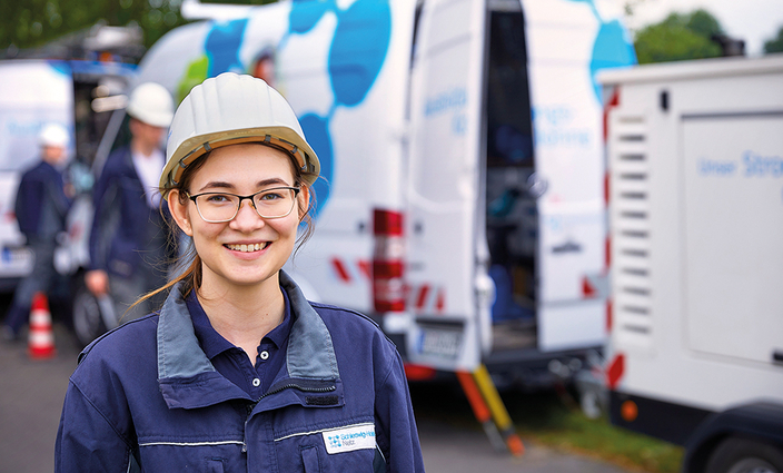 Junge Person in Arbeitskleidung mit Schutzhelm lächelt in die Kamera, im Hintergrund Fahrzeuge und zwei weitere Personen Junge Person in Arbeitskleidung mit Schutzhelm lächelt in die Kamera, im Hintergrund Fahrzeuge und zwei weitere Personen