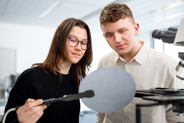 Zwei junge Personen stehen neben einem Mikroskop und begutachten eine Scheibe Zwei junge Personen stehen neben einem Mikroskop und begutachten eine Scheibe