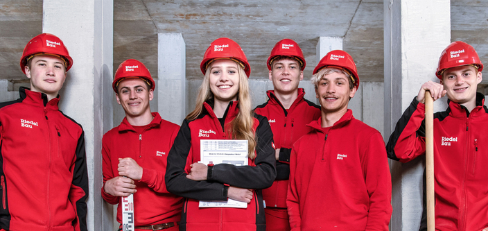 6 junge Mitarbeitende der Riedel Bau AG in roter Arbeitskleidung mit Helmen lächeln den Betrachter an.