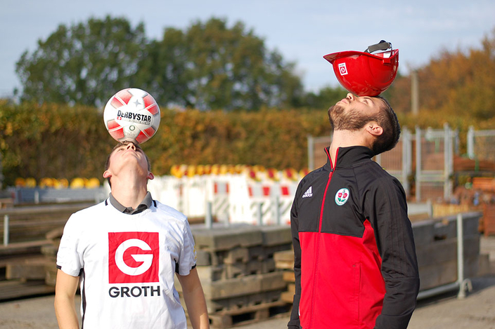 Zwei Personen, eine balanciert einen Fußball auf dem Kopf, ein anderer einen Bauhelm. Zwei Personen, eine balanciert einen Fußball auf dem Kopf, ein anderer einen Bauhelm.