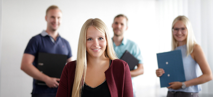 Junge Person mit langen Haaren lächelt in die Kamera, im Hintergrund drei weitere Personen mit Laptops und Unterlagen in der Hand Junge Person mit langen Haaren lächelt in die Kamera, im Hintergrund drei weitere Personen mit Laptops und Unterlagen in der Hand
