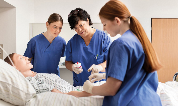 Drei Menschen in blauer Pflegebekleidung am Krankenbett, Auszubildende übt die Versorgung an einer Pflegepuppe