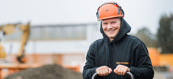 Junger Mensch im Kapuzenpulli mit Firmenlogo und Schutzhelm arbeitet mit einer Maschine auf der Baustelle