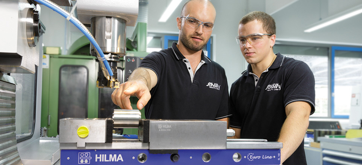 Zwei junge Menschen in Poloshirts mit Firmenlogo sehen sich eine Maschine in der Produktionsstätte an