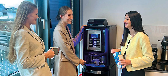 Drei junge lächelnde Personen im Gespräch am Kaffeeautomaten Drei junge lächelnde Personen im Gespräch am Kaffeeautomaten