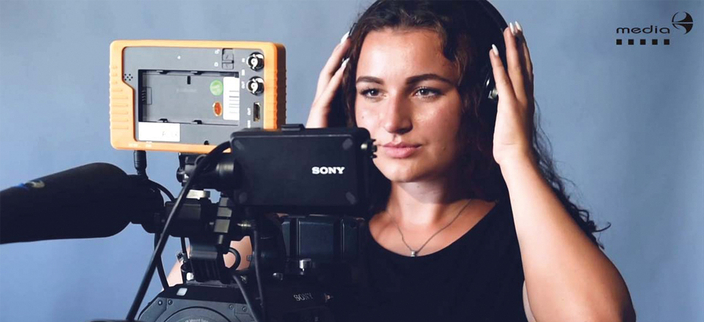 Eine Person steht hinter einer professionellen Videokamera. Sie trägt Kopfhörer und hält diese mit beiden Händen an den Ohren. Ihr Blick ist konzentriert zur Kamera gerichtet. Eine Person steht hinter einer professionellen Videokamera. Sie trägt Kopfhörer und hält diese mit beiden Händen an den Ohren. Ihr Blick ist konzentriert zur Kamera gerichtet.