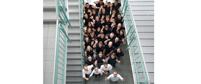 Eine Gruppe von etwa 40 Personen steht dicht beisammen in einem Treppenhaus und winkt nach oben zur Kamera. Alle tragen dunkle oder weiße Kleidung. Eine Gruppe von etwa 40 Personen steht dicht beisammen in einem Treppenhaus und winkt nach oben zur Kamera. Alle tragen dunkle oder weiße Kleidung.