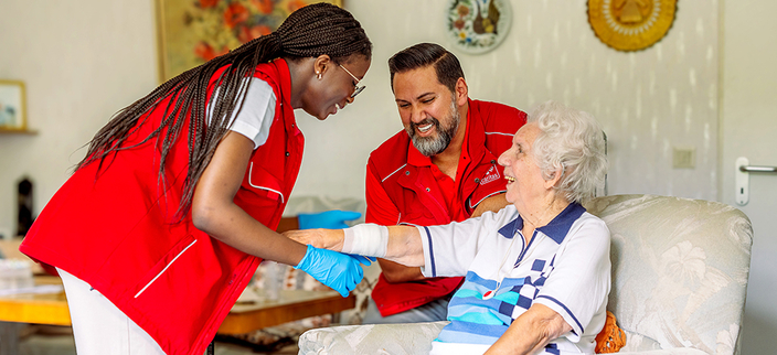Zwei Personen in roter Pflegebekleidung kontrollieren den Verband am Arm einer Seniorin Zwei Personen in roter Pflegebekleidung kontrollieren den Verband am Arm einer Seniorin