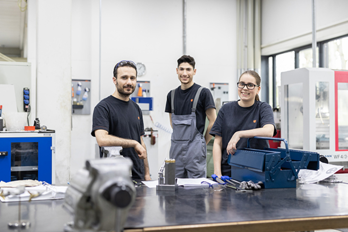 Drei Mitarbeitende in der Produktionsstätte lächeln in die Kamera Drei Mitarbeitende in der Produktionsstätte lächeln in die Kamera