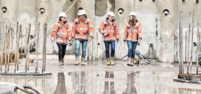 Vier Personen in orangefarbener Schutzkleidung und Helmen gehen lächelnd über eine nasse Baustelle. Vier Personen in orangefarbener Schutzkleidung und Helmen gehen lächelnd über eine nasse Baustelle.