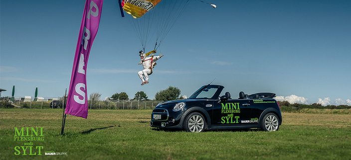 Ein Fallschirmspringer landet neben einem Auto mit der Aufschrift: Mini Flensburg meets Sylt Ein Fallschirmspringer landet neben einem Auto mit der Aufschrift: Mini Flensburg meets Sylt