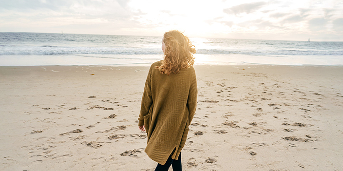 Person mit langen Haaren und Strickjacke geht im Sand am Meer entlang Person mit langen Haaren und Strickjacke geht im Sand am Meer entlang