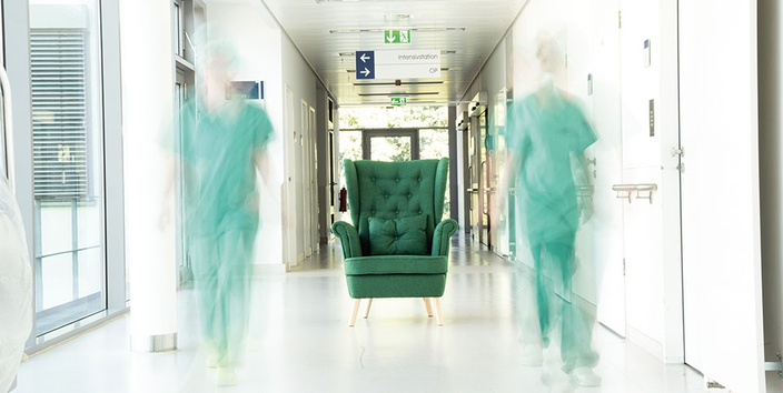 Flur in einem Krankenhaus, mitt steht ein grüner Ohrensessel, rechts und links daneben laufen zwei Pflegekräfte in grüner Bekleidung.