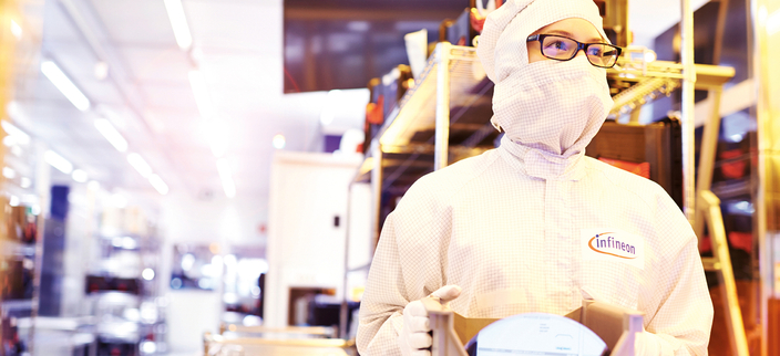 Eine Person in Reinraumkleidung steht in einem hell beleuchteten Labor und hält ein technisches Gerät in den Händen. Auf dem Anzug steht „Infineon“. Eine Person in Reinraumkleidung steht in einem hell beleuchteten Labor und hält ein technisches Gerät in den Händen. Auf dem Anzug steht „Infineon“.