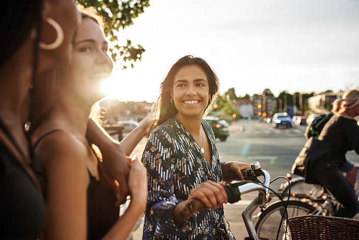 Junge Frau schiebt Fahrrad und unterhält sich mit anderen jungen Leuten Junge Frau schiebt Fahrrad und unterhält sich mit anderen jungen Leuten