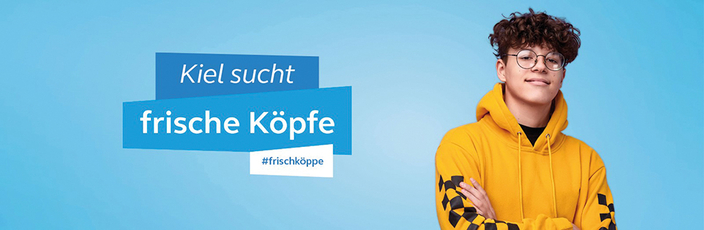 Kampagnenbild der Landeshauptstadt Kiel Junger Mann steht vor blauen Hintergrund Banner links daneben "Kiel sucht frische Köpfe" #frischeköppe