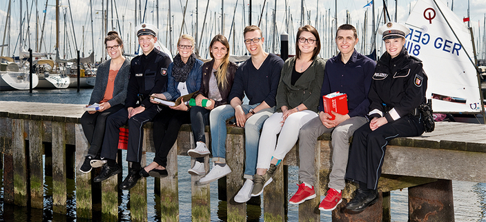 Acht Azubis aus verschiedenen Bereichen sitzen auf einem Steg, im Hintergrund Segelboote Acht Azubis aus verschiedenen Bereichen sitzen auf einem Steg, im Hintergrund Segelboote