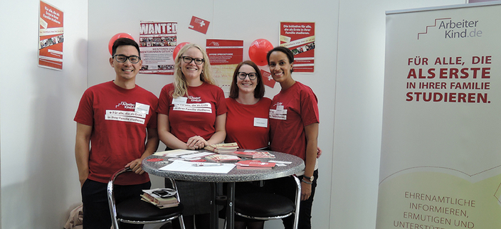 Vier Personen in roten T-Shirts stehen an einem Messetisch. Daneben steht ein Roll-Up von ArbeiterKind.de mit der Aufschrift: FÜR ALLE, DIE ALS ERSTE IN IHRER FAMILIE STUDIEREN. Vier Personen in roten T-Shirts stehen an einem Messetisch. Daneben steht ein Roll-Up von ArbeiterKind.de mit der Aufschrift: FÜR ALLE, DIE ALS ERSTE IN IHRER FAMILIE STUDIEREN.