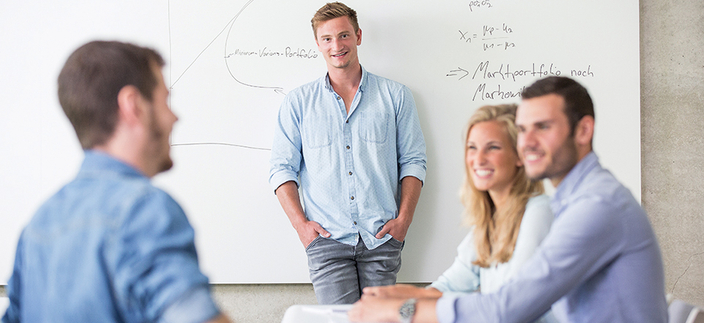 Eine Person steht vor einem Whiteboard mit Diagrammen und Formeln. Weitere drei Personen sitzen lächelnd an einem Tisch. Eine Person steht vor einem Whiteboard mit Diagrammen und Formeln. Weitere drei Personen sitzen lächelnd an einem Tisch.