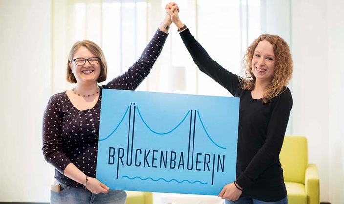 Zwei Frauen bauen mit den Armen eine Brücke und halten ein Schild "Brückenbauerin" Zwei Frauen bauen mit den Armen eine Brücke und halten ein Schild "Brückenbauerin"