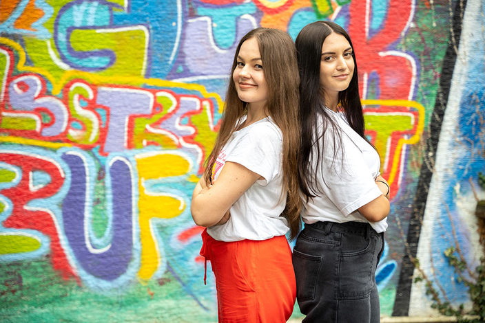 Zwei Personen stehen mit dem Rücken zueinander vor einer Wand mit Graffitis und blicken lächelnd in die Kamera. Zwei Personen stehen mit dem Rücken zueinander vor einer Wand mit Graffitis und blicken lächelnd in die Kamera.