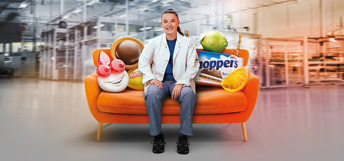 Junge Frau auf orange farbenen Sofa umgeben von übergroßen Storck Produkten Junge Frau auf orange farbenen Sofa umgeben von übergroßen Storck Produkten