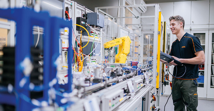 Eine Person steht in einem Technikraum und bedient eine Steuerungseinheit. Vor ihr befindet sich eine lange Werkbank mit vielen Kabeln, Geräten und einem gelben Roboterarm. Eine Person steht in einem Technikraum und bedient eine Steuerungseinheit. Vor ihr befindet sich eine lange Werkbank mit vielen Kabeln, Geräten und einem gelben Roboterarm.