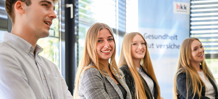 Vier junge Personen sitzen nebeneinander und lächeln in die Kamera, im Hintergrund ein Roll-Up der hkk Krankenkasse