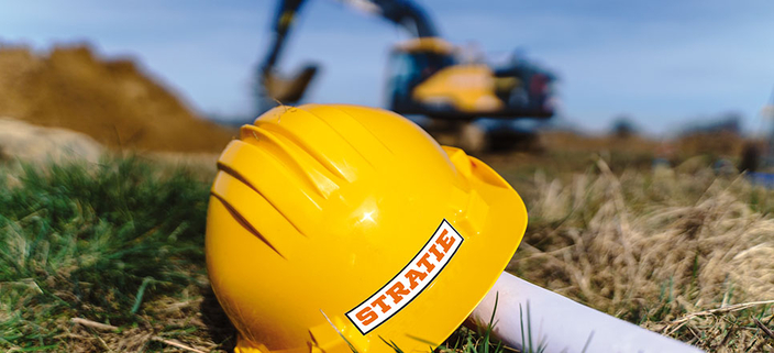 Bauhelm von STRATIE Bau GmbH liegt auf einer Baustelle im Gras, im Hintergrund ein Bagger bei der Arbeit Bauhelm von STRATIE Bau GmbH liegt auf einer Baustelle im Gras, im Hintergrund ein Bagger bei der Arbeit