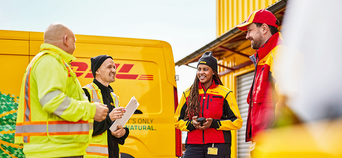4 Mitarbeitende, die sich am Verteilzentrum besprechen, im Hintergrund steht ein DHL Fahrzeug 4 Mitarbeitende, die sich am Verteilzentrum besprechen, im Hintergrund steht ein DHL Fahrzeug