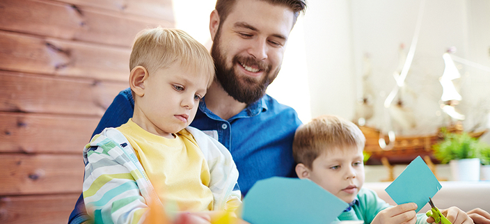Zwei Kinder sitzen auf dem Schoß einer erwachsenen Person und basteln mit Schere und Papier. Zwei Kinder sitzen auf dem Schoß einer erwachsenen Person und basteln mit Schere und Papier.