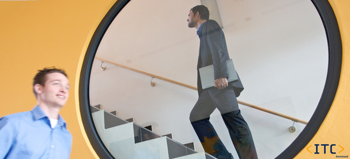 Junge lächelnde Person geht an einem kreisförmigen Fenster vorbei, durch das man eine Person im Anzug mit Laptop die Treppe hochgehen sieht Junge lächelnde Person geht an einem kreisförmigen Fenster vorbei, durch das man eine Person im Anzug mit Laptop die Treppe hochgehen sieht