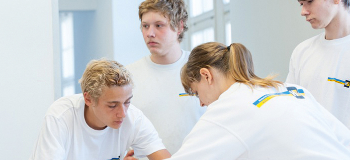 Vier junge Personen in weißen T-Shirts mit Firmenlogo arbeiten an einem Tisch Vier junge Personen in weißen T-Shirts mit Firmenlogo arbeiten an einem Tisch