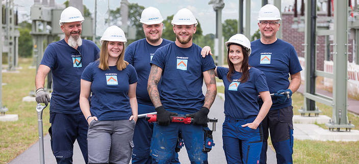 Sechs lächelnde Mitarbeitende mit Helm in blauen T-Shirts mit Firmenlogo an einem Umspannwerk Sechs lächelnde Mitarbeitende mit Helm in blauen T-Shirts mit Firmenlogo an einem Umspannwerk