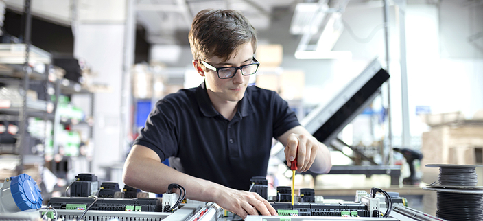 Eine Person mit Brille arbeitet konzentriert mit einem Schraubenzieher an einer offenen Steuerungseinheit. Eine Person mit Brille arbeitet konzentriert mit einem Schraubenzieher an einer offenen Steuerungseinheit.