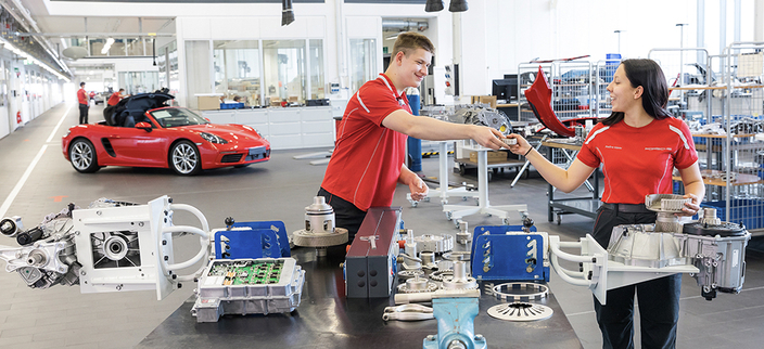 Zwei junge Menschen in roter Arbeitskleidung arbeiten lächelnd an einem Werkstatttisch mit Autoteilen. Im Hintergrund steht ein roter Sportwagen in einer modernen Werkhalle. Zwei junge Menschen in roter Arbeitskleidung arbeiten lächelnd an einem Werkstatttisch mit Autoteilen. Im Hintergrund steht ein roter Sportwagen in einer modernen Werkhalle.