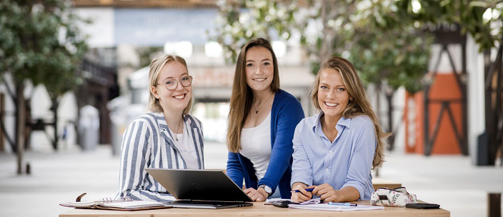Drei Studierende vor einem Laptop lächeln in die Kamera