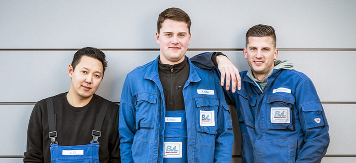 Drei lächelnde Mitarbeitende in blauer Arbeitskleidung vor einer grauen Wand