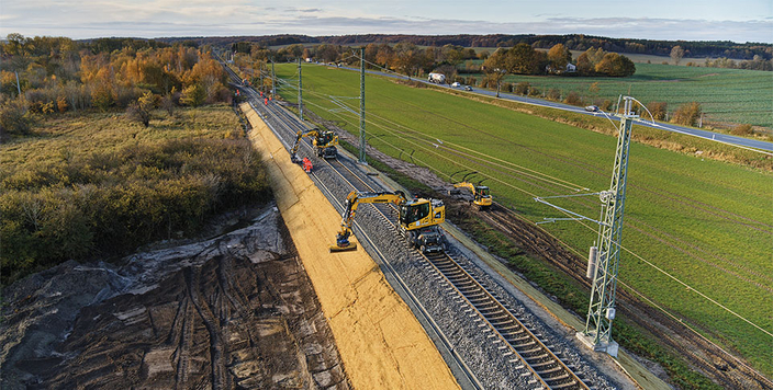 Mehrere Baufahrzeuge und Personen arbeiten an einer Bahnstrecke, die durch eine ländliche Landschaft mit Feldern und Wald führt. Mehrere Baufahrzeuge und Personen arbeiten an einer Bahnstrecke, die durch eine ländliche Landschaft mit Feldern und Wald führt.