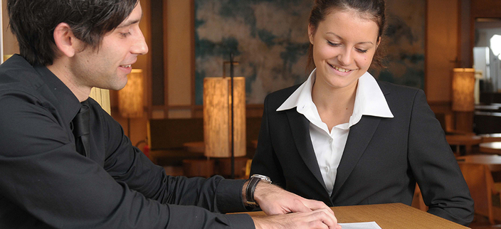 Lächelnde Person an einem Empfangstresen zeigt einer anderen Person etwas auf einem Papier Lächelnde Person an einem Empfangstresen zeigt einer anderen Person etwas auf einem Papier