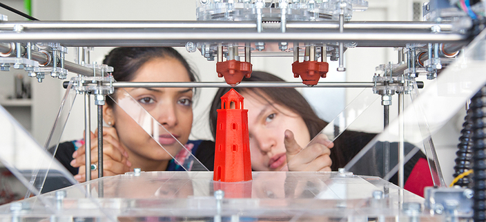 Großaufnahme eines 3D Druckers, beim Drucken eines roten Turmes im Hintergrund zwei Mitarbeiterinnen, die sich darüber austauschen Großaufnahme eines 3D Druckers, beim Drucken eines roten Turmes im Hintergrund zwei Mitarbeiterinnen, die sich darüber austauschen