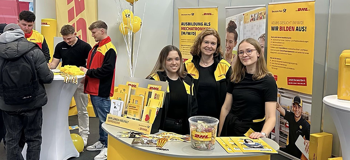 Drei lächelnde Personen am Messetresen der DHL Group – Deutsche Post, im Hintergrund ein Stehtisch mit vier weitern Personen Drei lächelnde Personen am Messetresen der DHL Group – Deutsche Post, im Hintergrund ein Stehtisch mit vier weitern Personen