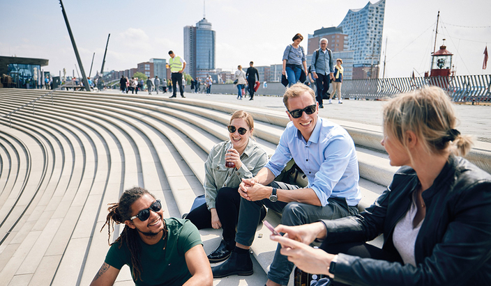 Vier junge Menschen sitzen auf einer Treppe an den Landungsbrücken, im Hintergrund die Elbphilharmonie Vier junge Menschen sitzen auf einer Treppe an den Landungsbrücken, im Hintergrund die Elbphilharmonie