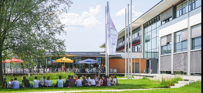 Vor dem Gebäude der Hochschule Hof sitzen viele Personen im Rasen. Vor dem Gebäude der Hochschule Hof sitzen viele Personen im Rasen.
